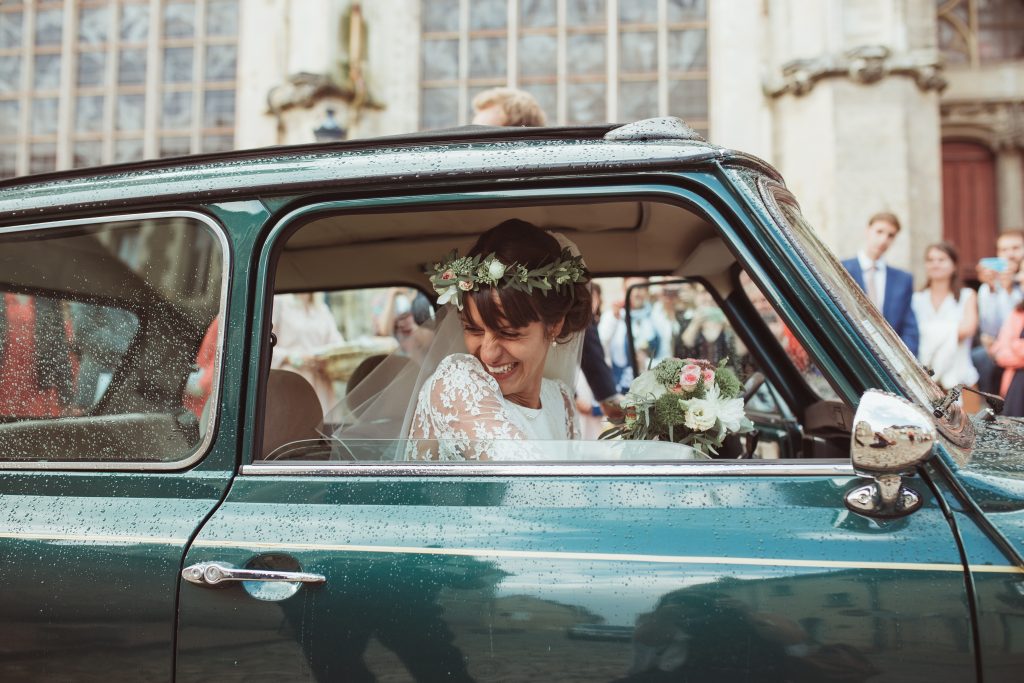 La mariée riant à l'arrière d'une voiture ancienne verte devant la Cathédrale de Nevers, sous la pluie